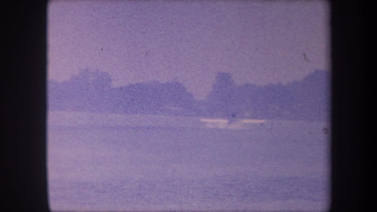 1959 California home-movie frame showing the same calm lake and distant shoreline; the small boat remains visible mid-water under a hazy sky.