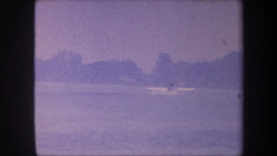 1959 California home-movie frame showing the same calm lake and distant shoreline; the small boat remains visible mid-water under a hazy sky.