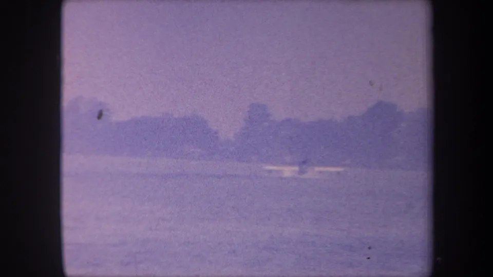 1959 California home-movie frame; the camera has drifted slightly, revealing more water surface, with a small white boat still visible against the purple-cast haze.