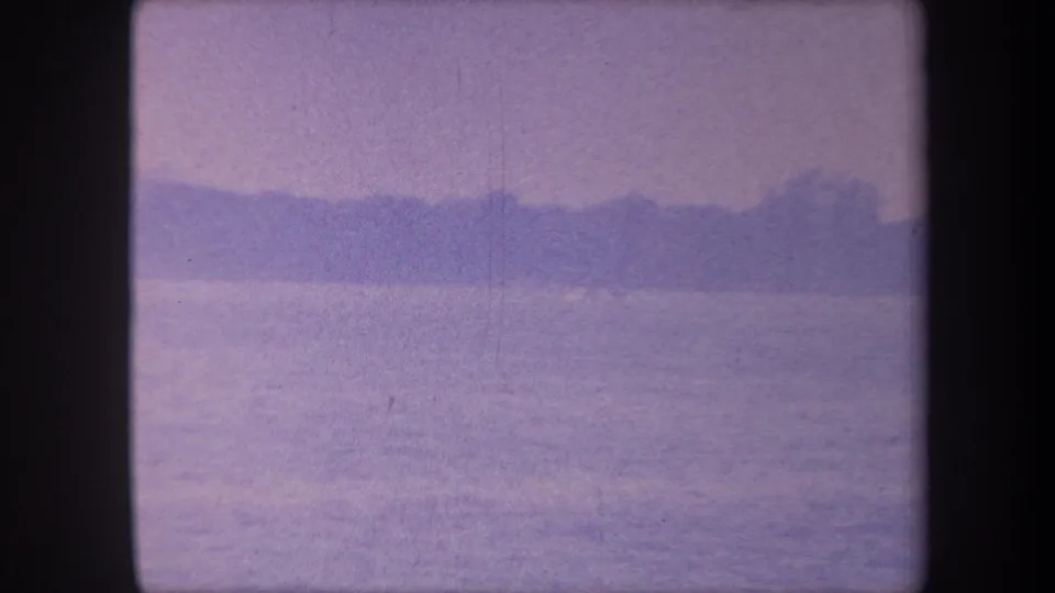 1959 8mm home movie frame of a still lake at Yucaipa, California, with a distant dark landmass silhouette under a lavender-cast sky.