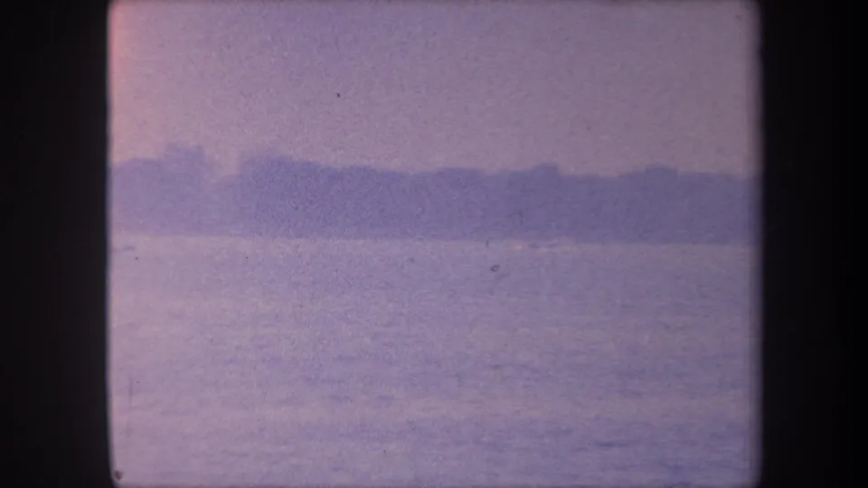 1959 8mm home movie frame with notable warm pink-lavender color shift, still lake water, and a dark silhouette landmass on the far horizon, Yucaipa, California.
