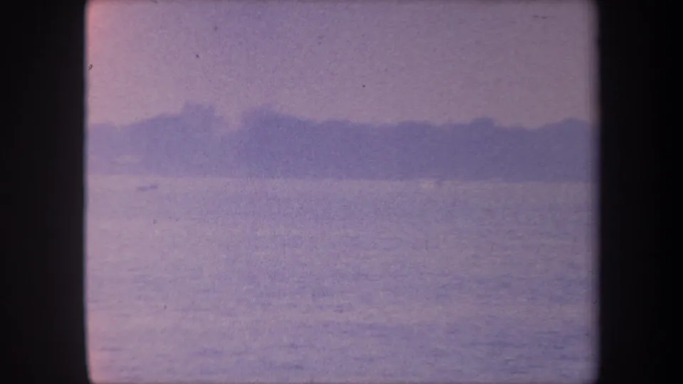 1959 8mm home movie frame of a calm California lake with a distant dark shoreline under a warm-toned lavender sky, with pronounced film-aging color and grain.