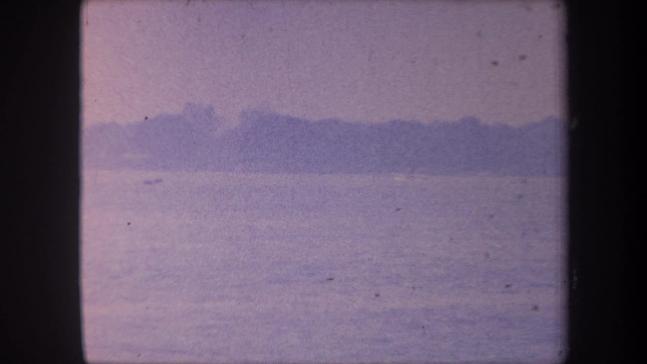 1959 California 8mm home movie frame — calm water expanse, distant treeline silhouette, heavy blue-purple color cast from aged emulsion.