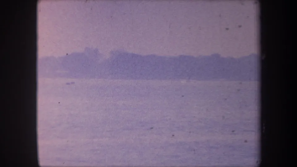 1959 California 8mm home movie frame — calm water expanse, distant treeline silhouette, heavy blue-purple color cast from aged emulsion.
