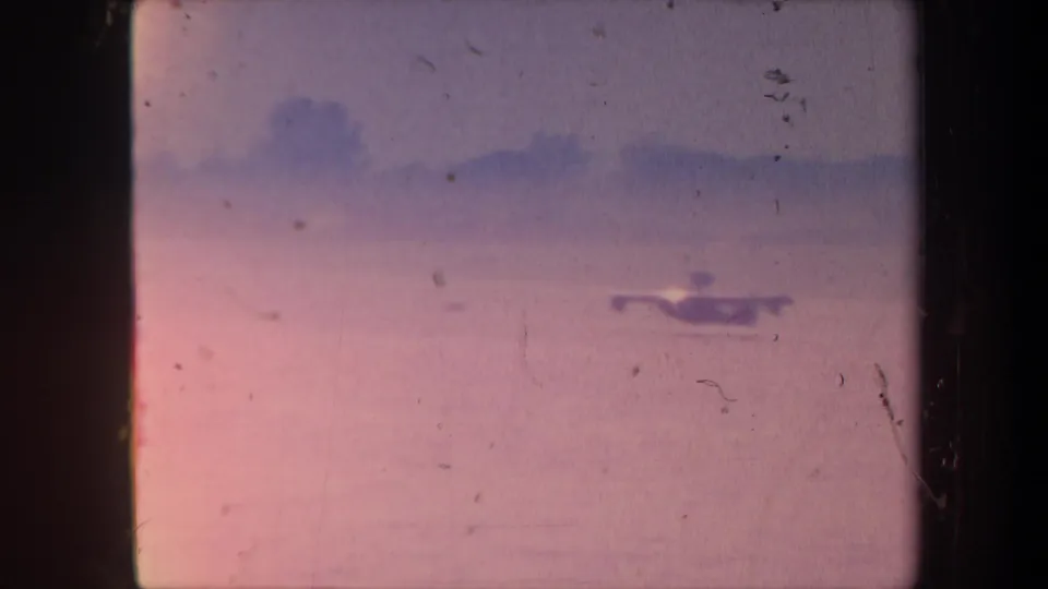 1959 California 8mm home movie — warm pink light-leak over water, small unidentified bright object on the water surface, distant shore silhouette above.
