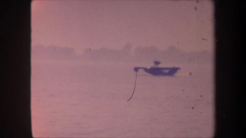 Small seaplane over or on a California lake surface in pink-shifted 1959 8mm footage; a diagonal film-scratch hair crosses the frame.