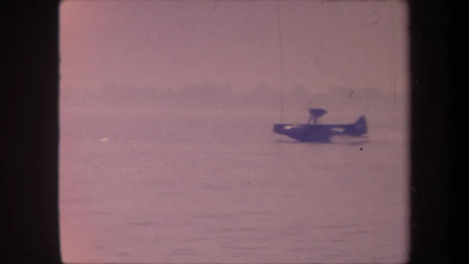 Small seaplane settled on the water surface of a California lake in 1959 8mm footage; relatively clean frame with visible water surface and distant shore.