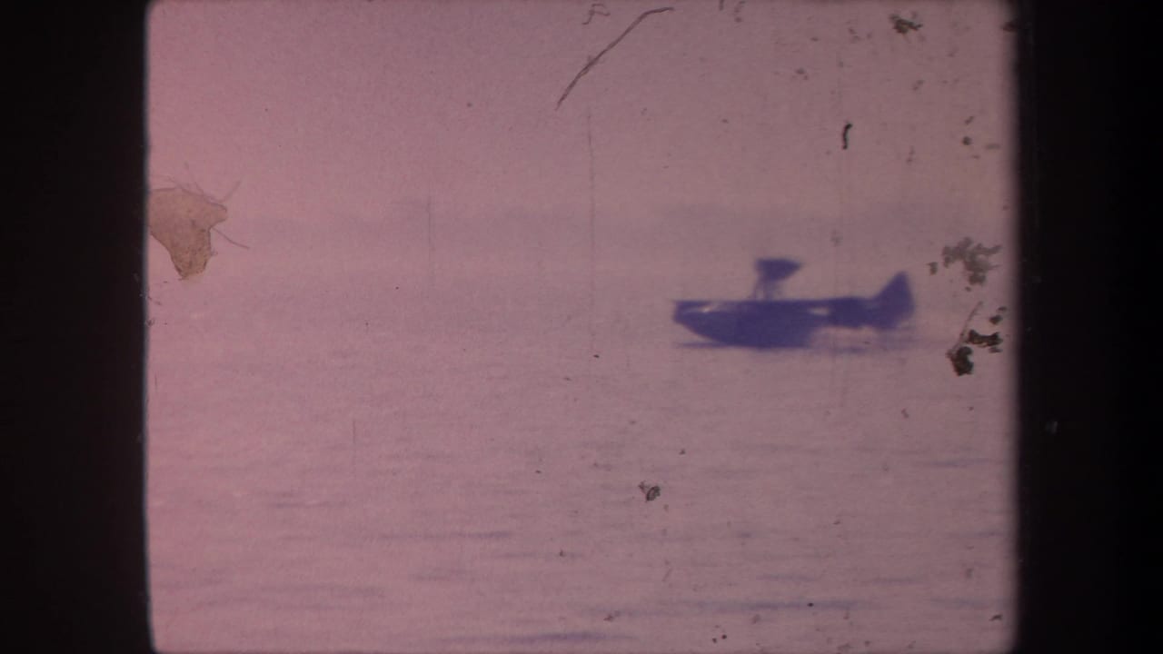 Small dark floatplane on open water, pinkish aged 8mm film, emulsion damage visible at upper frame edge