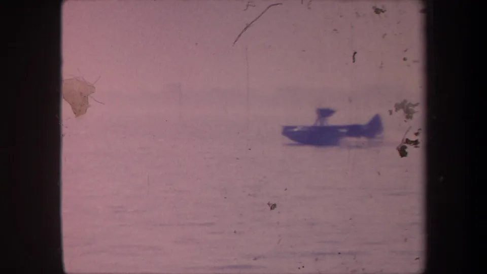 Dark-hulled aquaplane with visible pontoons on calm lake water, 1959 8mm home movie from Yucaipa, California, pinkish color cast and emulsion damage