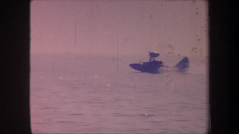 Dark-hulled aquaplane on calm lake surface, 1959 8mm home movie, visible film scratches and lavender color shift throughout frame