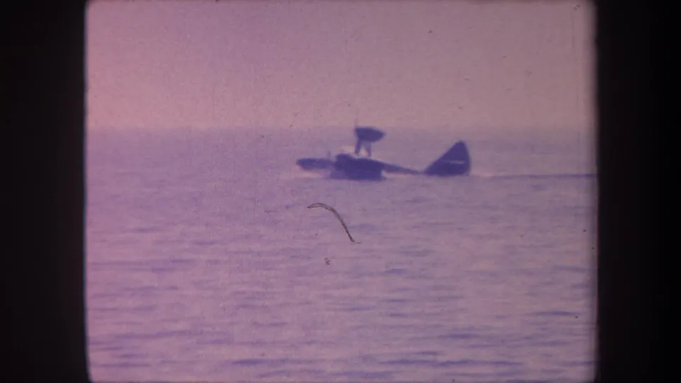 Small seaplane on open water with a prominent curved film scratch artifact in the foreground, 8mm home movie circa 1959
