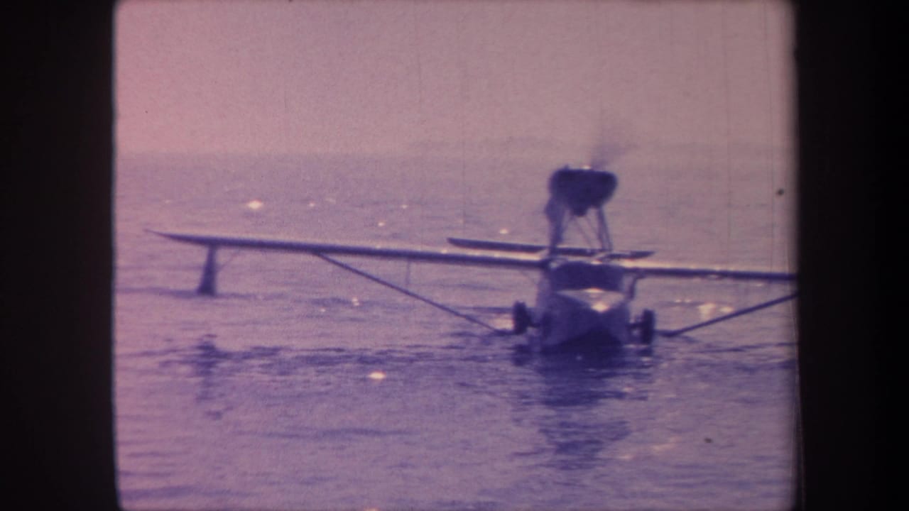 Amphibian aircraft on calm water, rear three-quarter view, propeller spinning with motion blur, strong purple color cast on 8mm film.