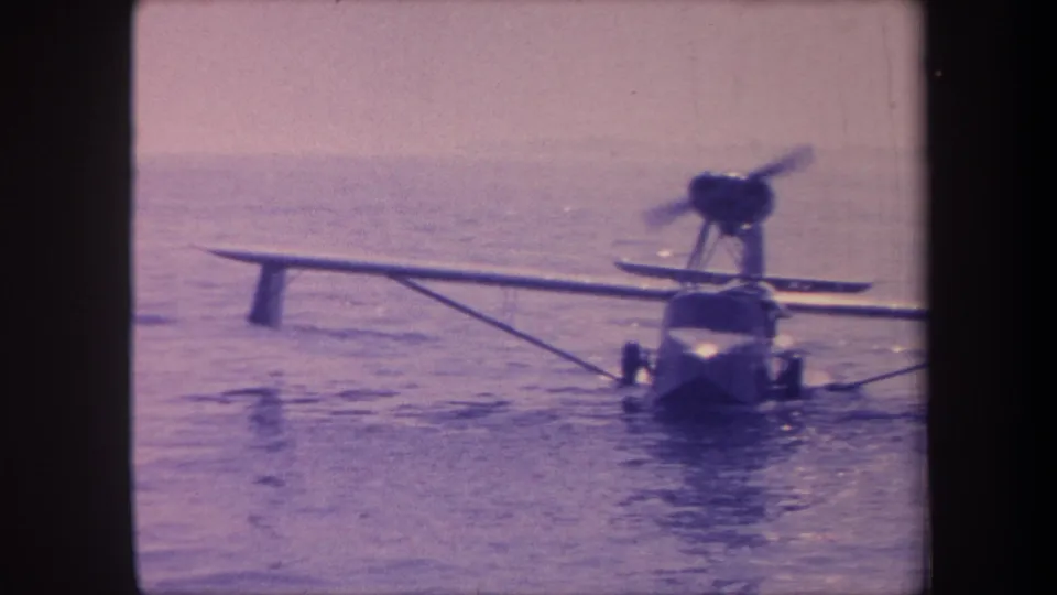 Single-engine amphibian aircraft floating on open water with a faint distant shoreline on the horizon, captured on 8mm film circa 1959 with pronounced purple color shift.