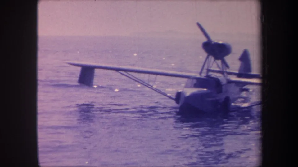 Low-angle view of a single-engine amphibian aircraft resting on water, hull and undercarriage prominent, shot on 8mm film circa 1959 with characteristic mid-century grain.