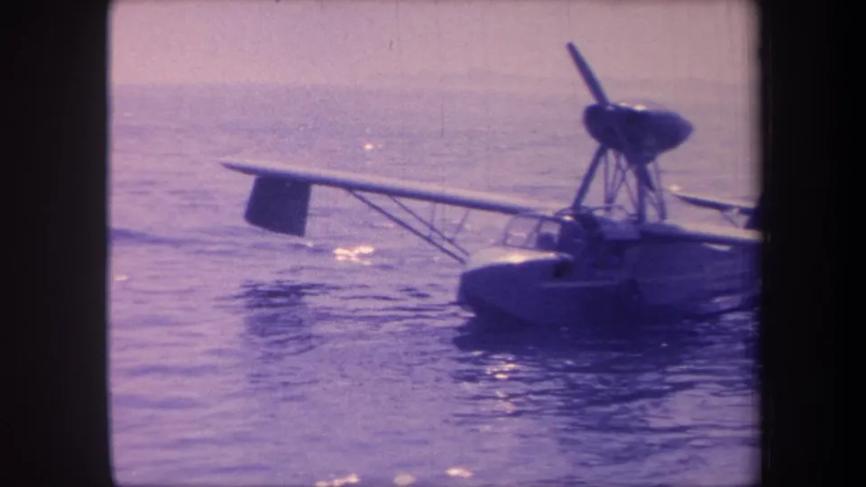 Right-side view of a single-engine amphibian aircraft floating on calm water, with strut and hull detail visible, shot on 8mm film circa 1959 with a strong purple color cast.