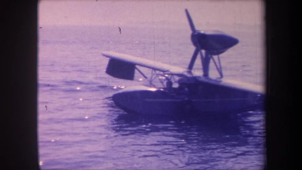 1959 8mm footage of a small flying-boat amphibian taxiing slowly on a California lake; strong purple-blue dye shift and consistent film grain throughout.