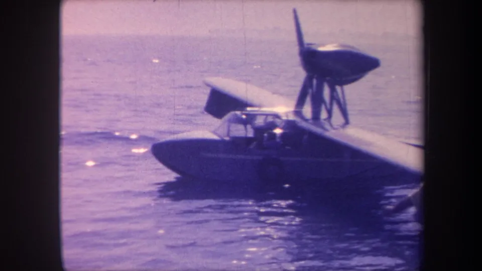 1959 8mm frame of a homebuilt flying-boat at closer range; a figure is visible near the cockpit; California lake setting; strong purple-blue color cast.