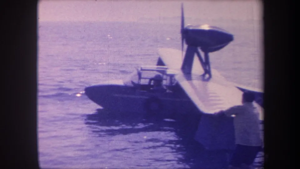 1959 8mm frame of a small flying-boat amphibian at close range; an observer is partially visible at lower right; California lake setting implied by metadata.