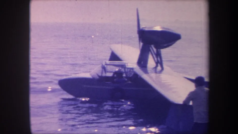 8mm home-movie frame, c. 1959, showing a single-engine flying boat on open water in California, person partially visible at right edge of frame.