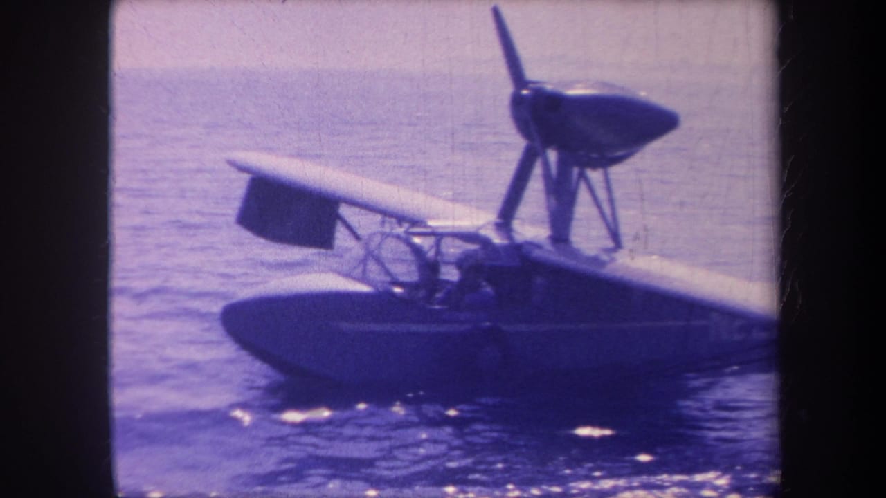 Small flying-boat seaplane resting on open water, starboard side view, pusher propeller on pylon struts above fuselage, open cockpit visible.