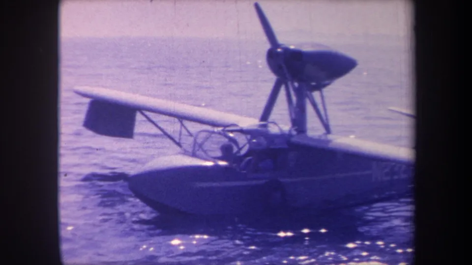 8mm home-movie frame circa 1959 showing the starboard side of a small flying-boat seaplane with a figure in the open cockpit, on calm coastal water.
