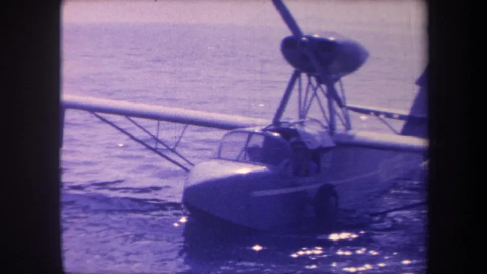 8mm home-movie frame circa 1959 showing a small flying-boat seaplane on calm water; shifted propeller blade position across frames suggests slow rotation.