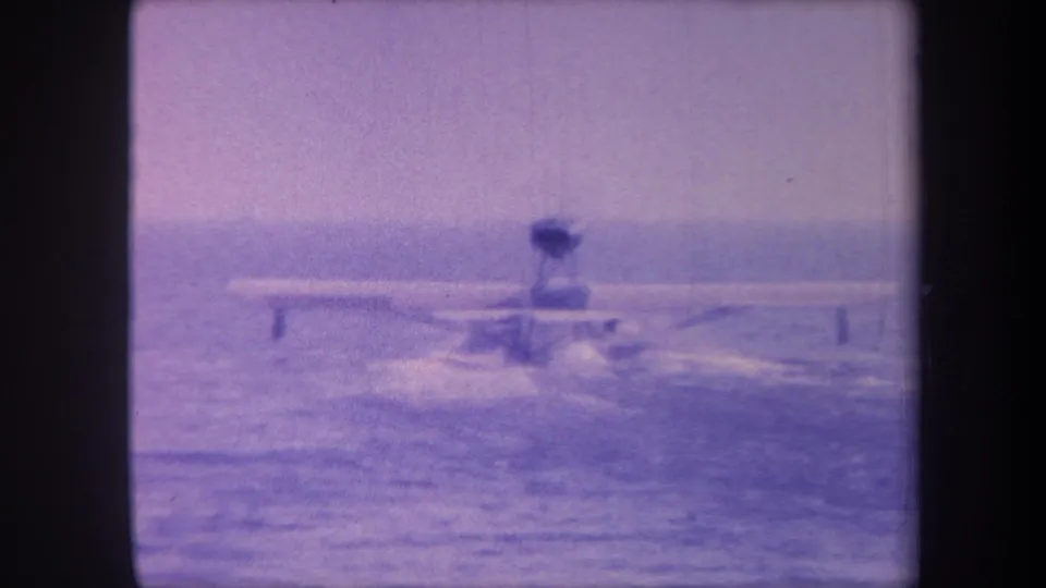 Seaplane on water takeoff run in circa 1959 8mm home movie footage; a person visible in the open cockpit as the aircraft builds speed across open water beneath a hazy sky.