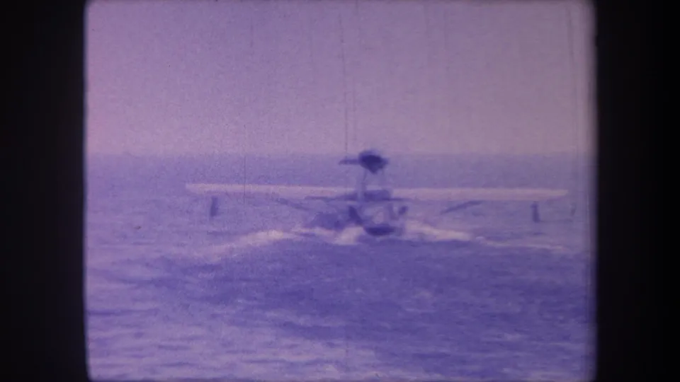 Seaplane mid-run on open water in circa 1959 8mm home movie; a person remains visible on the aircraft as it accelerates across calm water toward an indistinct horizon.