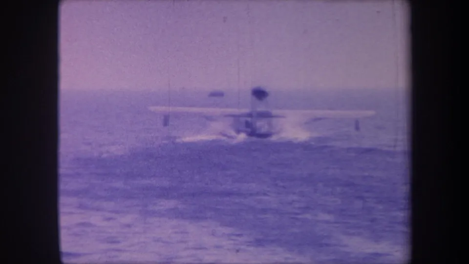 Final frame of a circa 1959 8mm seaplane clip; aircraft accelerating across open water with a figure visible on board, under a hazy sky with heavy blue-purple dye fading.