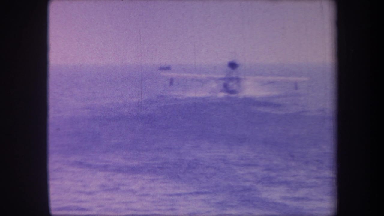 Seaplane on ocean surface mid-sequence, slightly repositioned, purple-tinted 8mm with grain and vignetting.