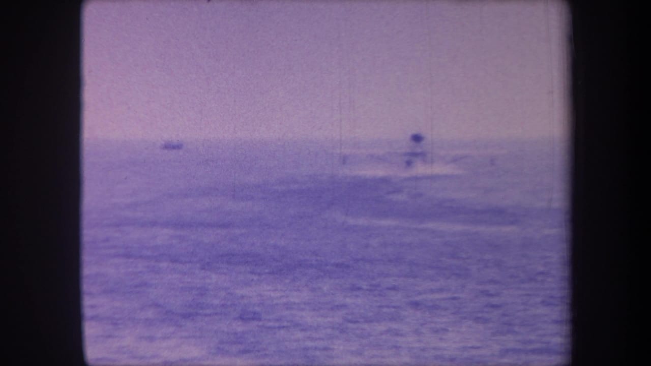 8mm home movie frame circa 1959 showing a distant motorboat on open sea, heavy magenta/violet dye-shift coloration and a faint second vessel on the far-left horizon.