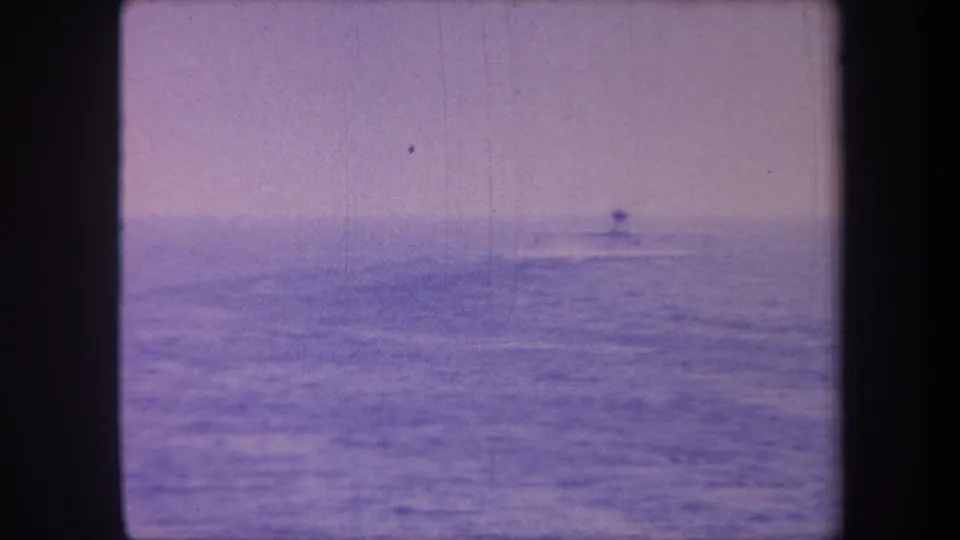 8mm home movie frame circa 1959 showing a lone boat silhouette on open sea; lavender dye-shift and grain are consistent with physically aged 8mm color film.
