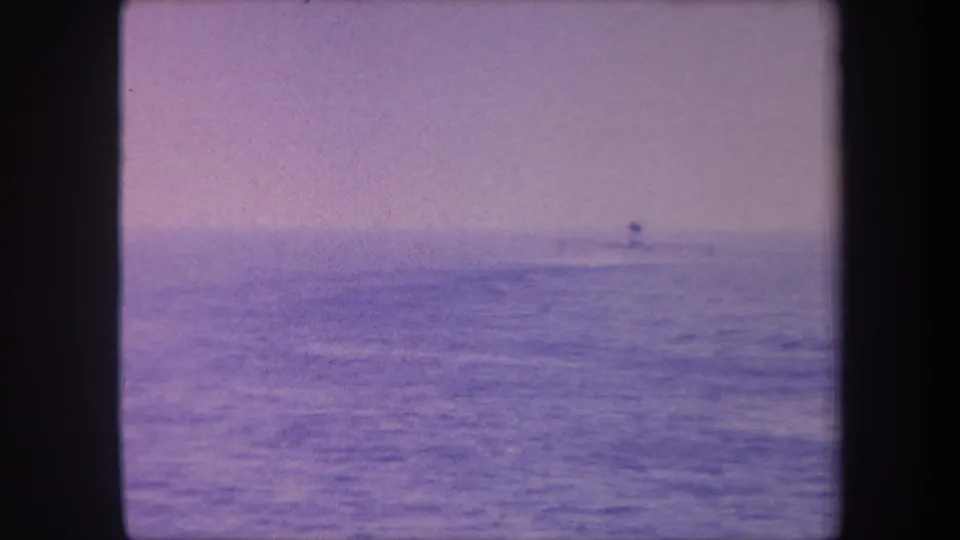 8mm home movie frame circa 1959 showing a tiny distant vessel on open sea; strong colour shift and grain mark this as a physically aged film original.