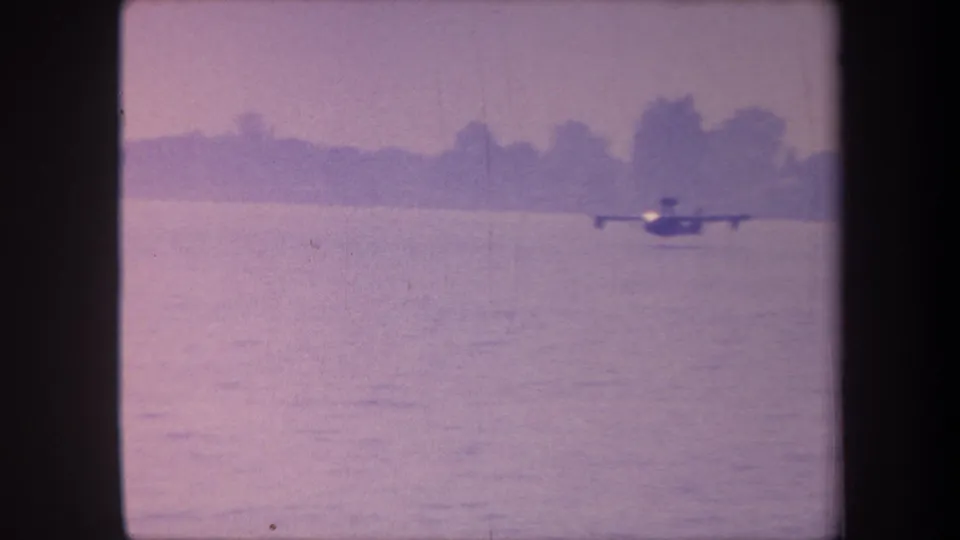 Flying-boat silhouette crossing calm water with low treeline horizon; color shifted to pink-lavender from dye fading, 8mm home movie circa 1959 California.