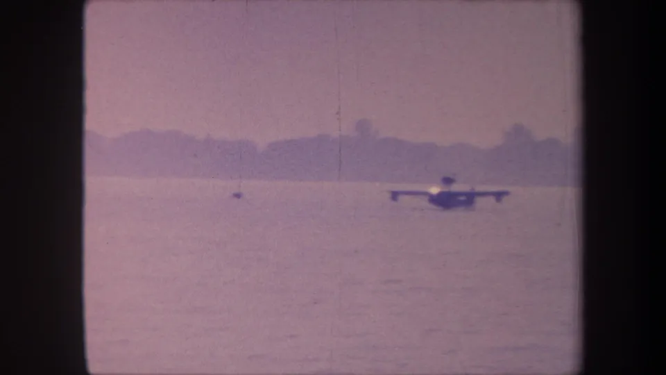 Flying boat or floatplane crossing calm water against a silhouetted treeline and hillside; 8mm home movie with characteristic pink dye fade, circa 1959 California.