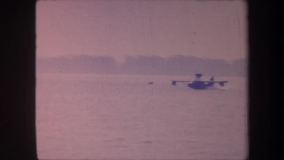Flying boat moving across calm water with low hillside treeline; warm pink color shift from aged 8mm emulsion, circa 1959 California.