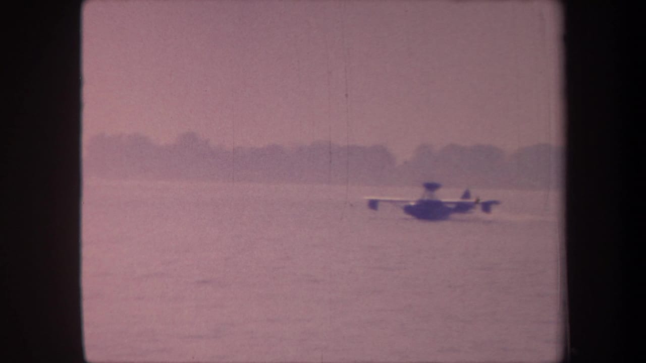 Floatplane taxiing on open lake with distant treeline on horizon, pink-shifted 8mm film frame.