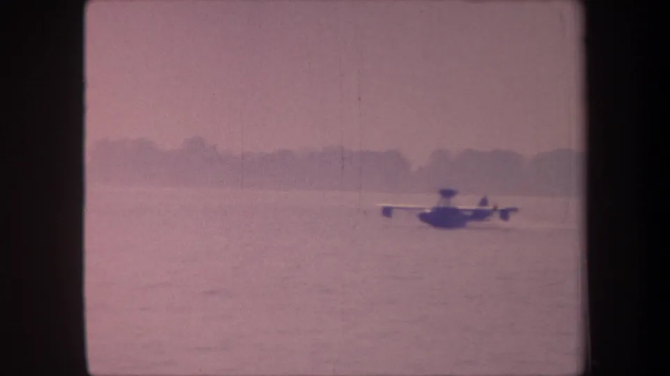 A floatplane moves across a calm lake surface in this 1959 8mm home-movie frame; heavy pink-magenta dye-shift from aged film stock.