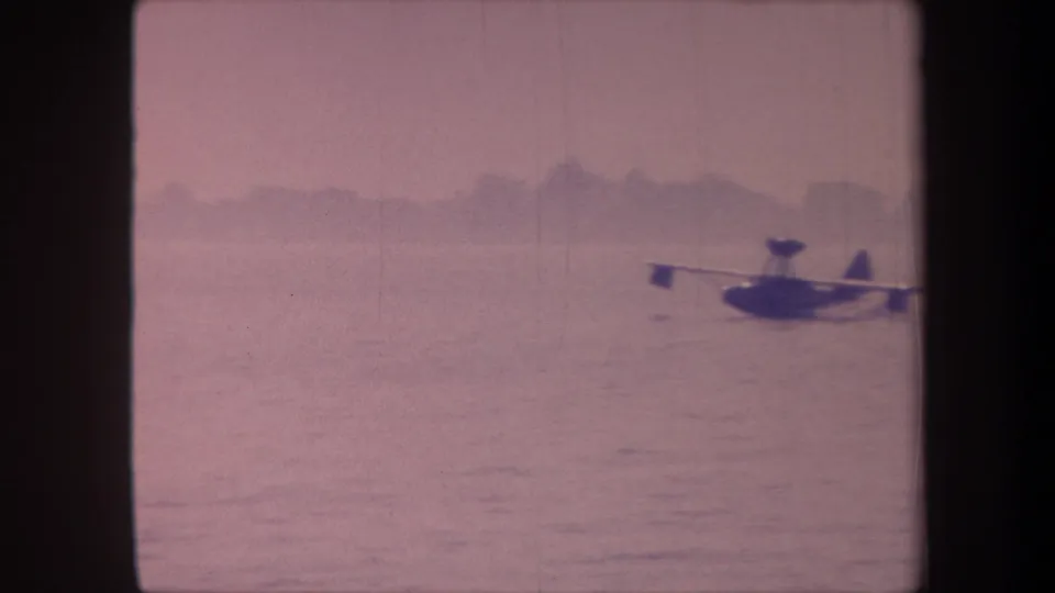 Floatplane moving across lake surface with low hills visible on the horizon; 1959 8mm home-movie footage with characteristic pink-magenta film aging.