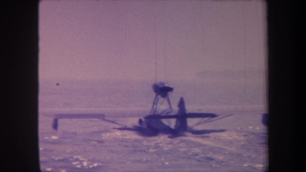 A high-wing propeller floatplane moves across a lake surface in California, circa 1959, captured on aged 8mm film with pronounced purple dye shift.