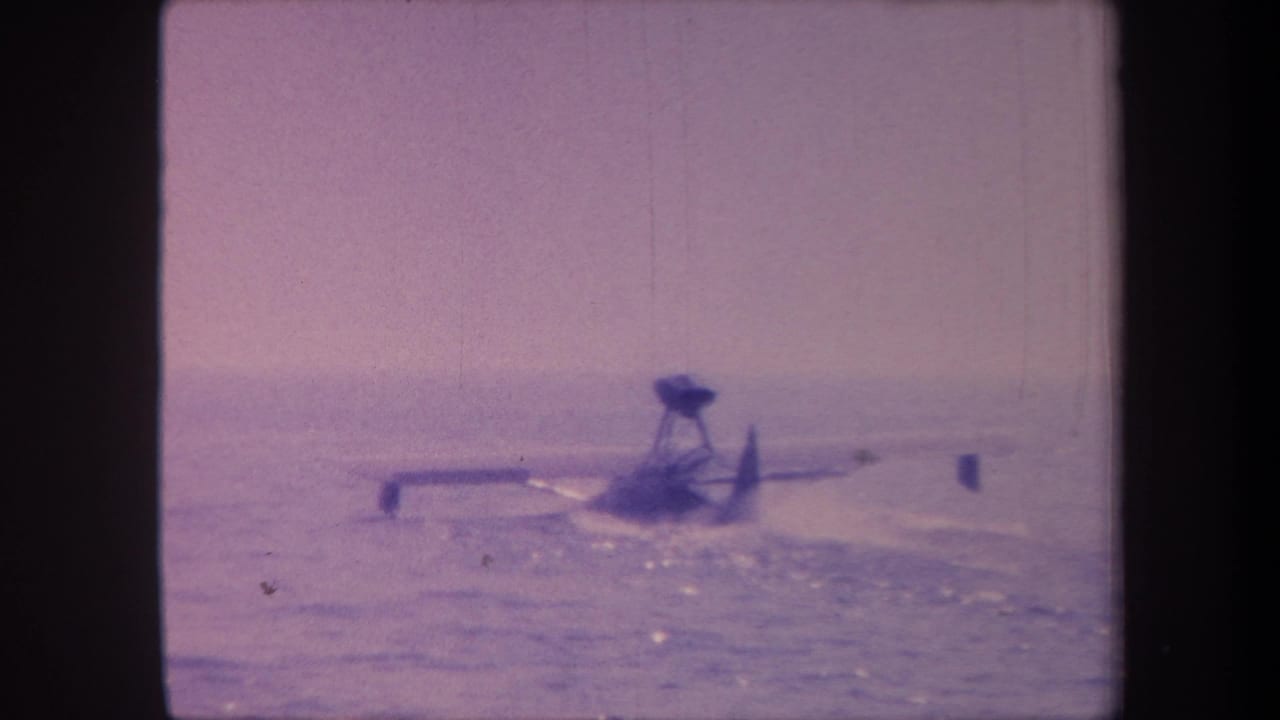 Floatplane on water surface in 1959 8mm home movie footage, purple-shifted film stock, hazy horizon and open water visible.