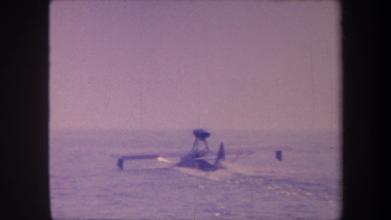 Floatplane continuing water takeoff run, purple-cast 8mm film, small buoy or marker visible at right of frame.