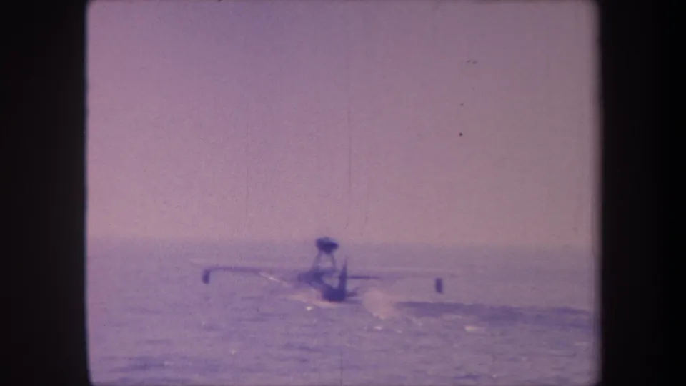 Floatplane at late stage of water takeoff in 1959 8mm home movie, characteristic purple film shift, water wake visible, open hazy sky.
