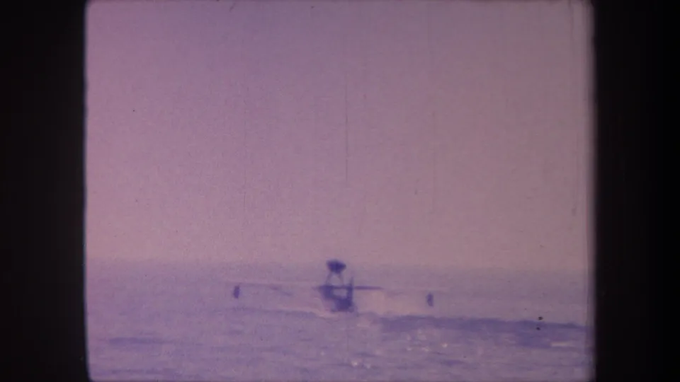 8mm home movie frame c. 1959 showing a hydroplane at mid-distance on open water with possible course markers and aged purple-lavender film cast.