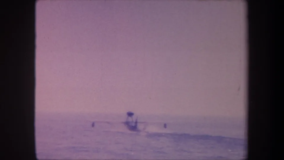 8mm home movie frame c. 1959 showing a hydroplane boat at distance on open water, sky dominating the upper half, aged purple-lavender film cast.