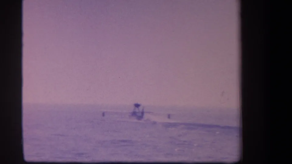 8mm home movie frame c. 1959 showing a hydroplane as a tiny silhouette near the waterline, lavender-purple sky dominating, characteristic of aged film stock.
