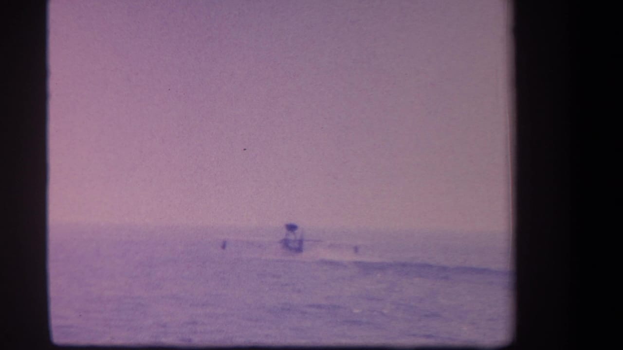 Lone surfer as small distant figure on open ocean, purple-tinted 8mm film with flat horizon and hazy sky.