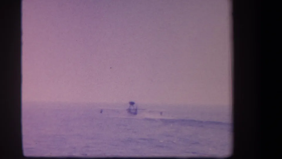 Distant surfer on open ocean in heavily purple-shifted 8mm home movie frame, likely California coast, circa 1959.