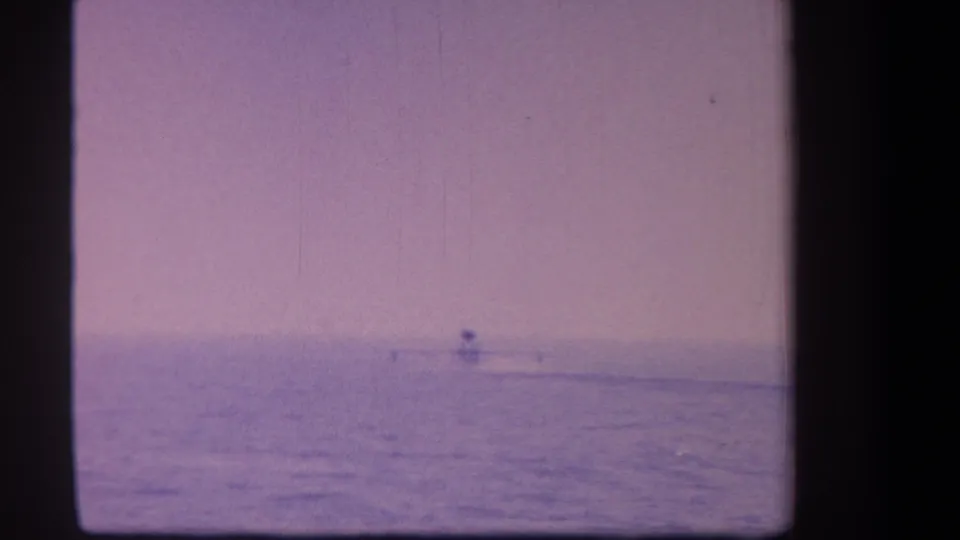 Surfer silhouette on broad ocean surface with heavy purple dye shift and grain; 8mm home movie, circa 1959.
