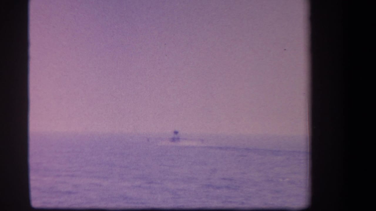 1959 8mm frame showing a small distant aircraft near the horizon over open water, with a lavender dusk cast.
